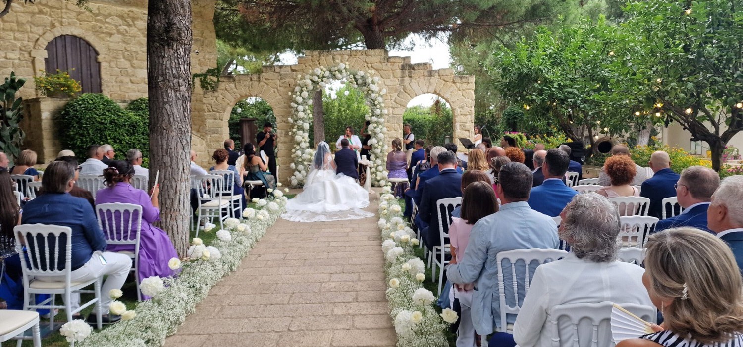Cerimonia all'aperto: la coppia davanti a un arco di fiori bianchi tra archi in pietra, gli invitati seduti ai lati del viale