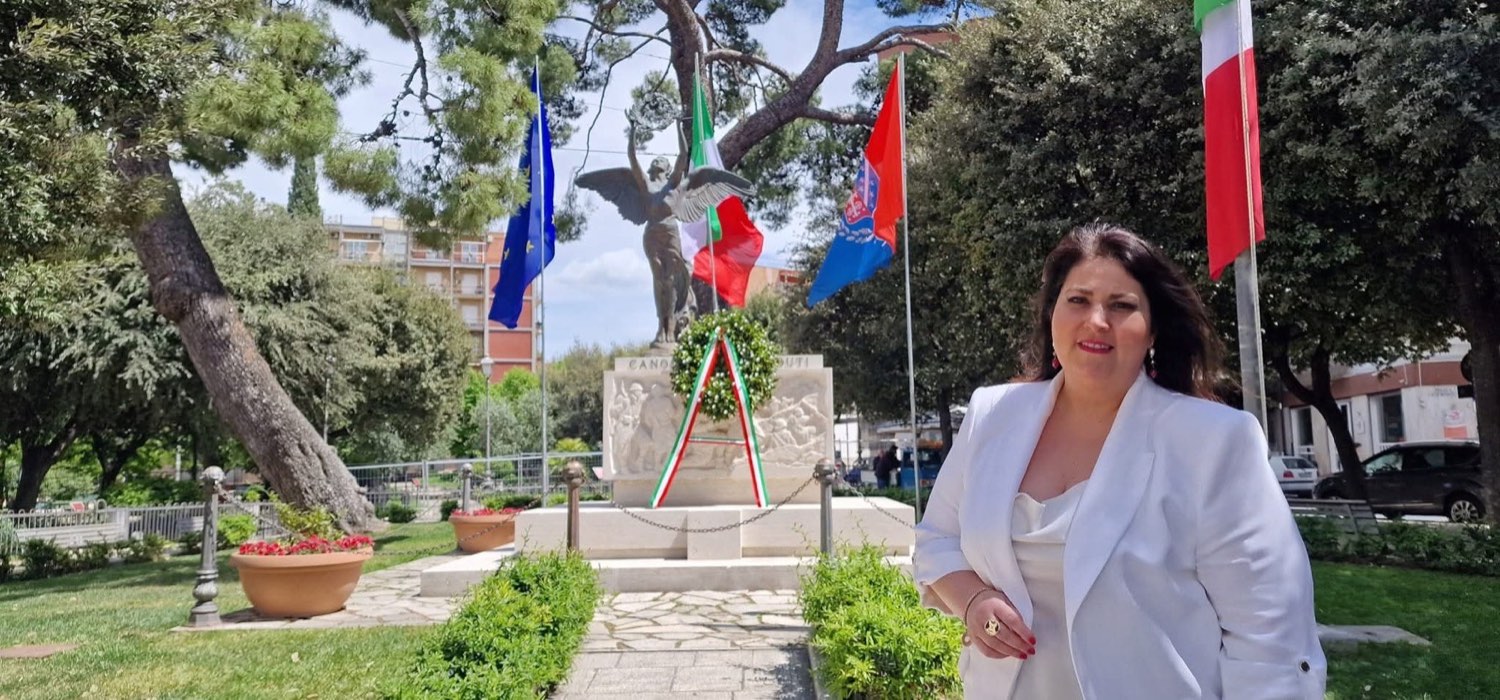 Claudia Vitrani accanto a un monumento ai caduti con corona d'alloro tricolore e bandiere